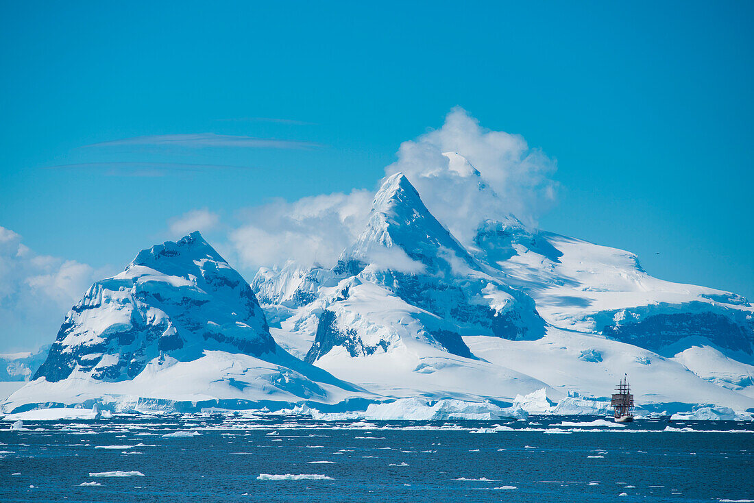 Sailing bark Europa amidst ice floes and icebergs with snow-covered mountains behind