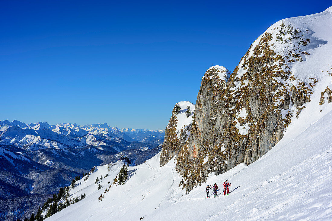 Three persons backcountry skiing ascending beneath rockface towa, Bavarian Alps in background, Rotwand, Spitzing, Bavarian Alps, Upper Bavaria, Bavaria, Germany