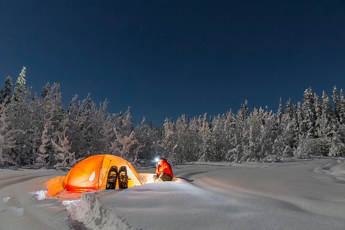Man with headlamp sits by a lit tent near an evergreen and snow covered forest, Gakona, Southcentral Alaska, Winter