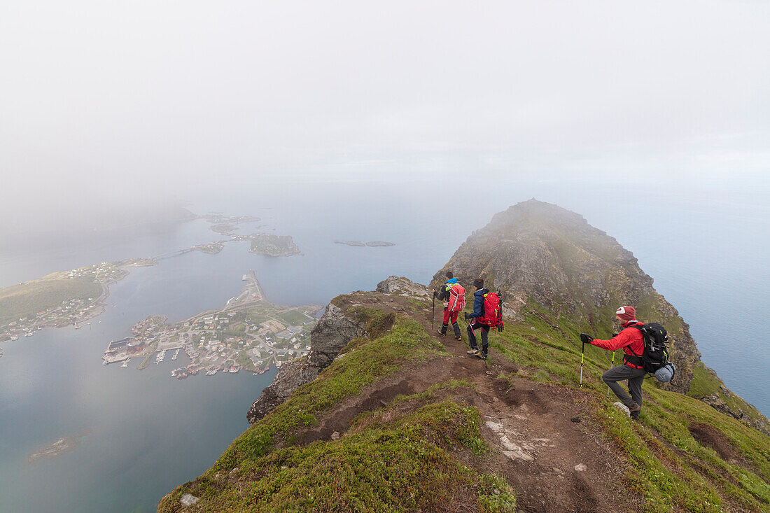 Hikers on top of rocky peak admire the blue sea surrounded by mist, Reinebringen, Moskenesoya, Lofoten Islands, Norway, Scandinavia, Europe