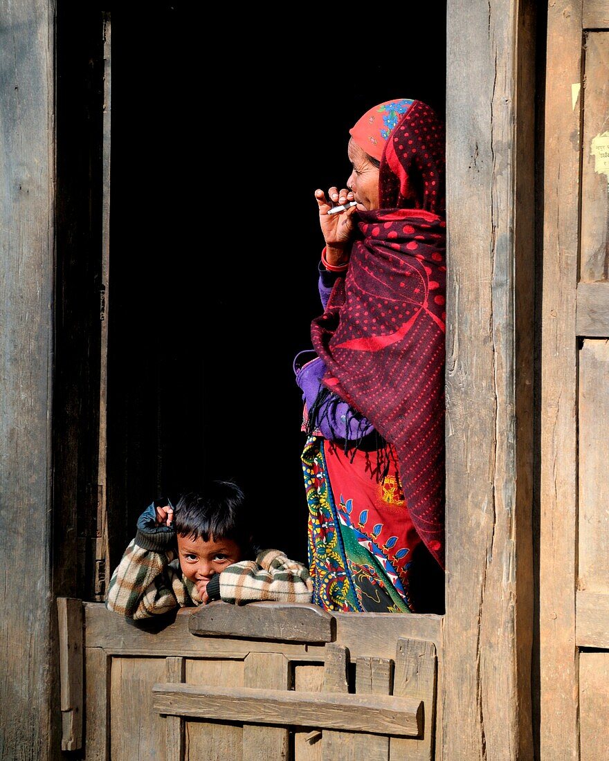 A mother looking at the horizon while smoking and a child who spy on passers, by and play with them with his smiling eyes, Nepal, Asia