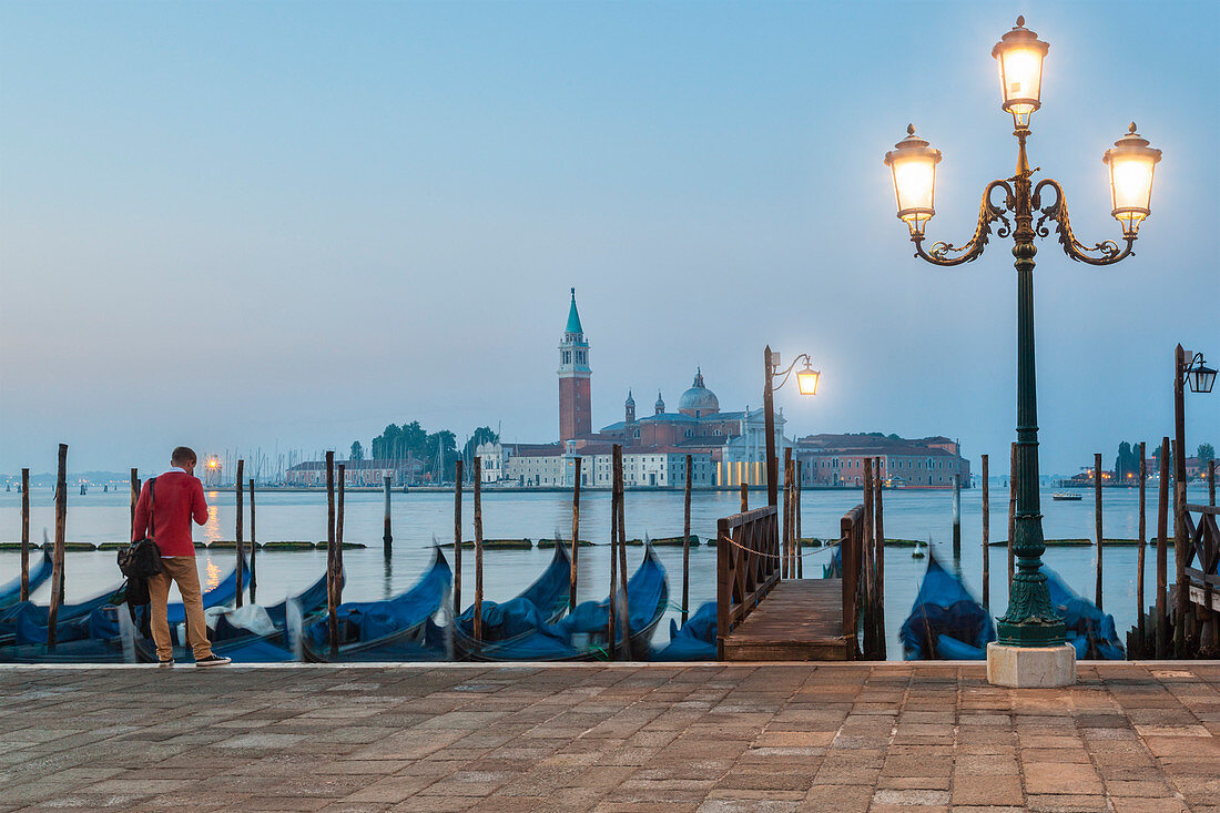 A solitary man is waiting in front of the gondolas docked along the Riva degli Schiavoni, in the background the island of San Giorgio Maggiore, Venice, Italy