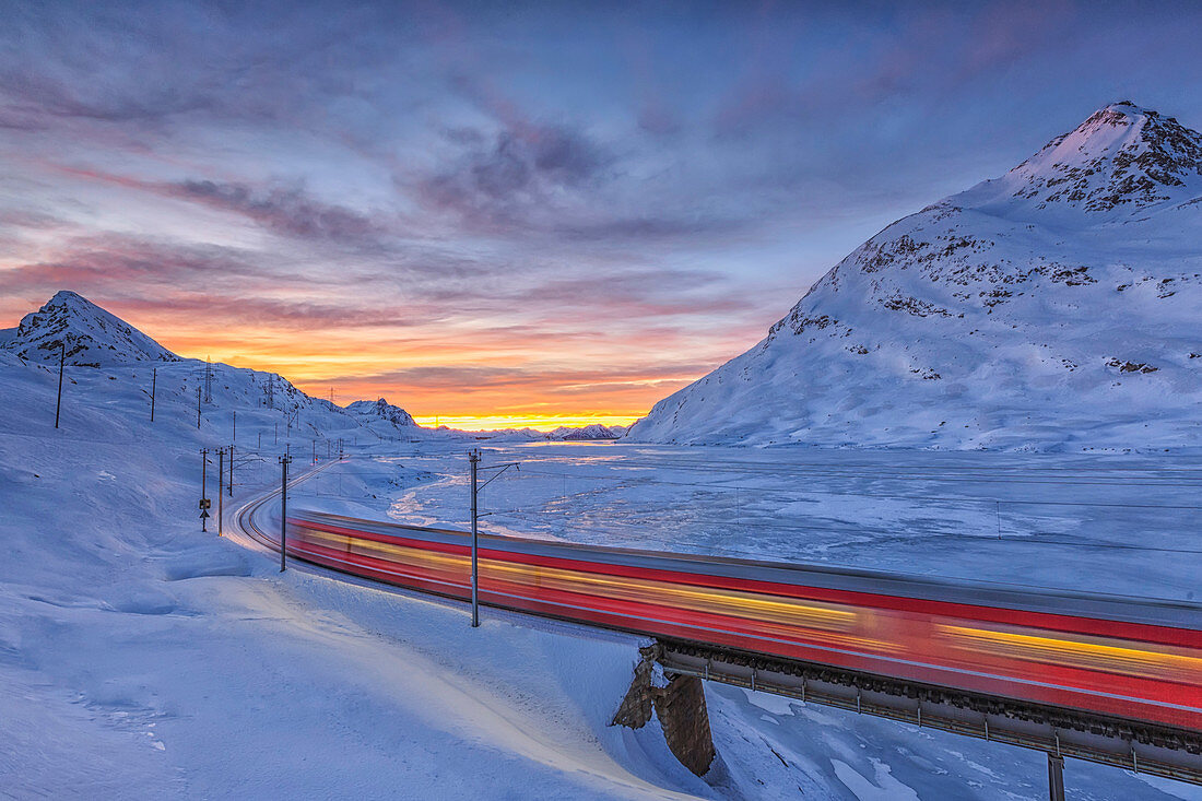 The Bernina Express passes on the shores of Lake Bianco completely frozen before the sun rises, Canton of Graubunden, Engadine, Switzerland, Europe