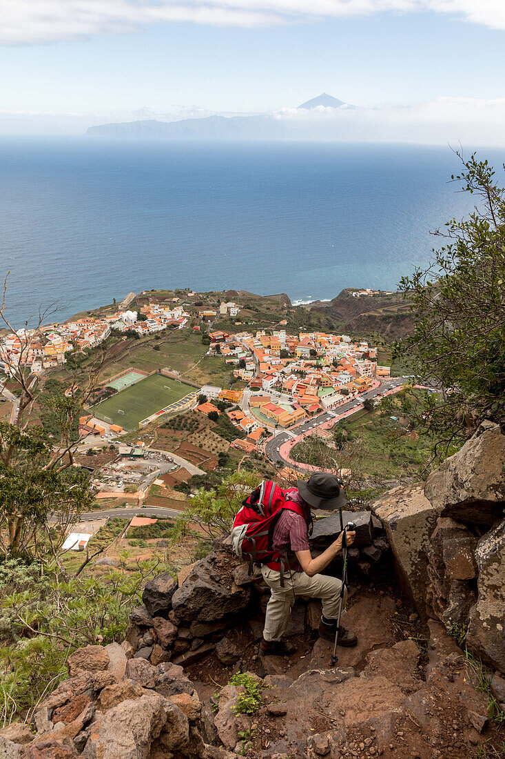 Wanderin mit rotem Rucksack, Blick Wanderweg zum Mirador de Abrante, Steilwand, in der Ferne der Pico del Teide, Teyde, höchste Berg Spaniens, Insel Teneriffa, unten liegt Agulo auf La Gomera, Kanaren, Kanarische , Inseln, Spanien