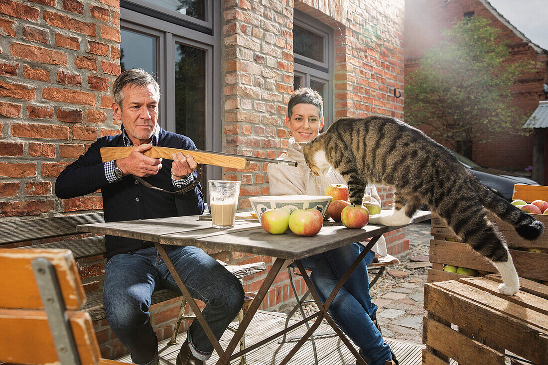 Playful man aiming toy rifle on tabby cat while sitting with woman in back yard