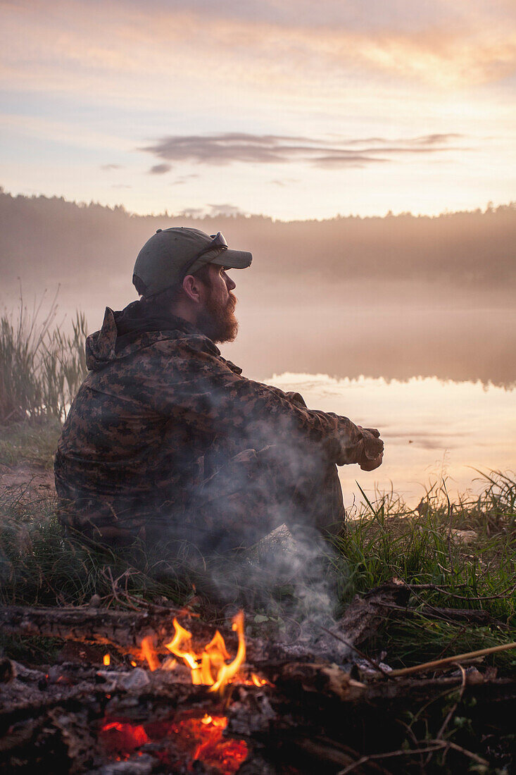 Side view of hunter sitting by bonfire at lakeshore during sunset