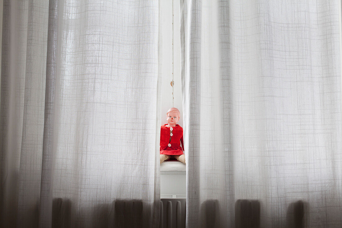 Doll sitting on windowsill behind curtains