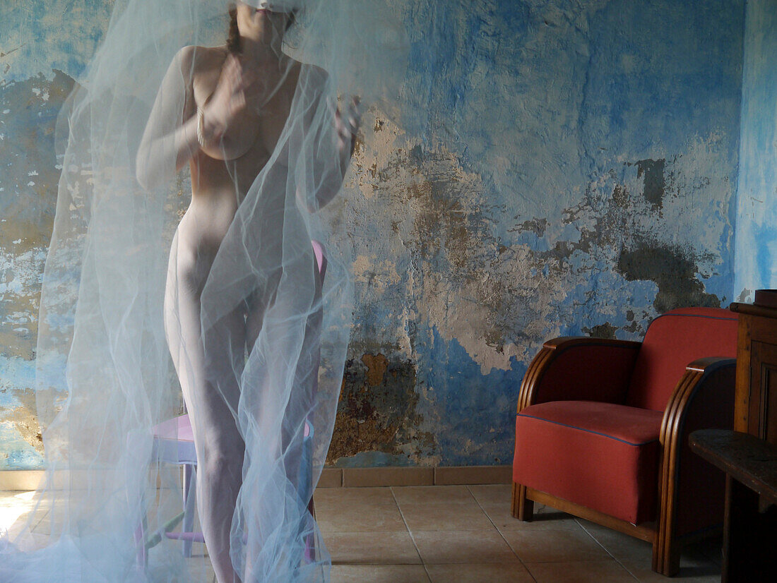 Front view of a female nude moving covered with a veil next to a red armchair