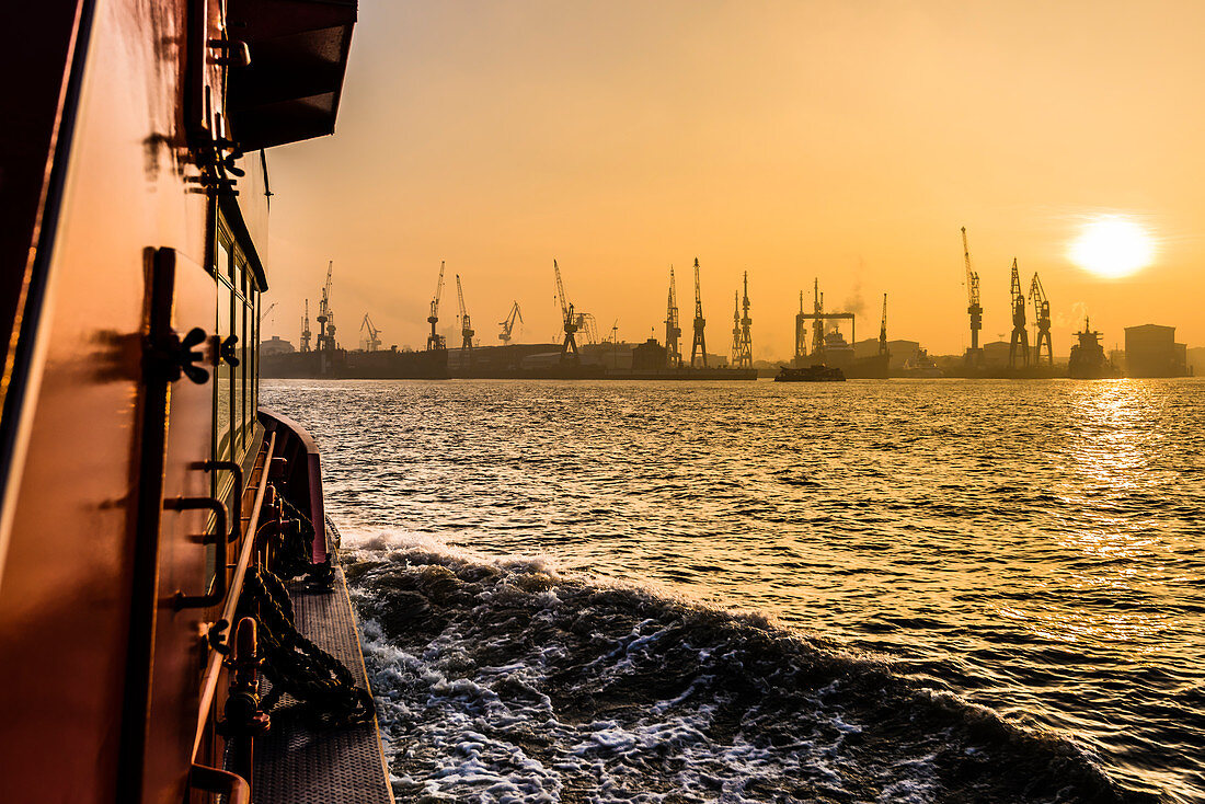Sonnenaufgang im Hamburger Hafen mit Blick auf Hafenanlagen und Kräne von der Elbfähre aus gesehen, Hamburg, Deutschland