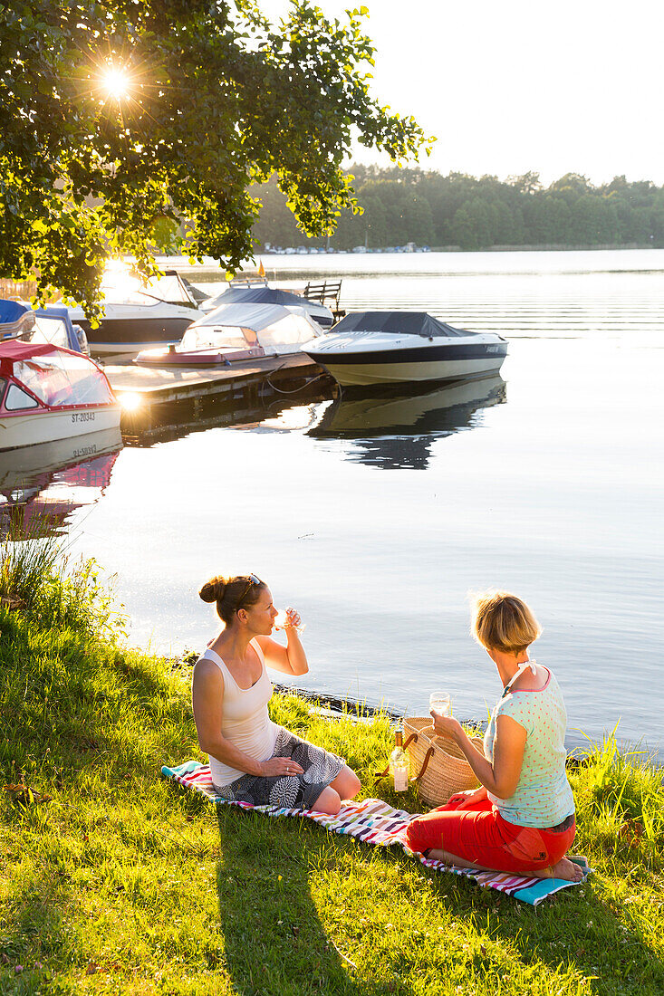 Two women drinking wine at the lakeshore, sunset, boats, lake Zethner See, summer, Mecklenburg lakes, Mecklenburg lake district, MR, Diemitz, Mecklenburg-West Pomerania, Germany, Europe