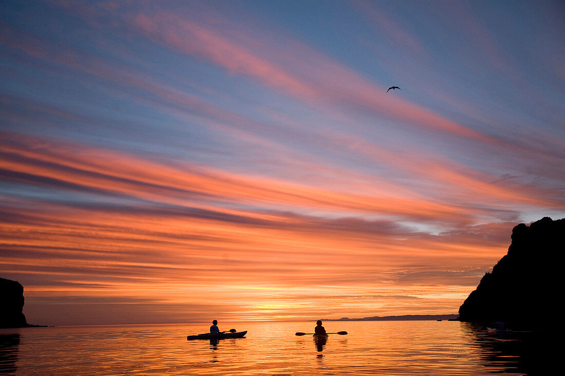 Loreto, Baja California Sur, Mexico. A couple relax in their sea kayaks watching an amazing orange blue sky at sunrise off the coast of Loreto, Baja California Sur, Mexico.