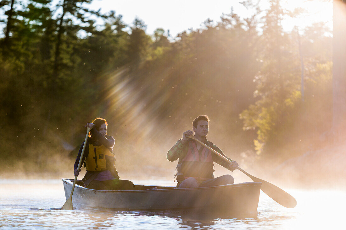 A Young Couple Paddles A Canoe Through Morning Mist On Long Pond In Maine's North Woods