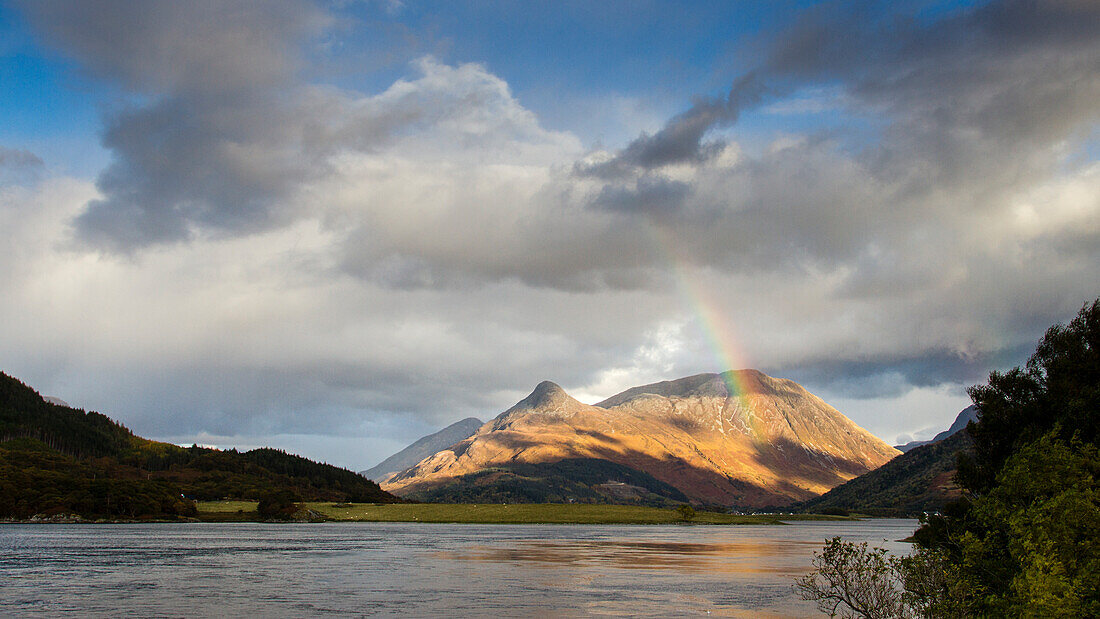 Scenic View Of A Lake And A Rainbow Cutting Through The Partly Cloudy Sky