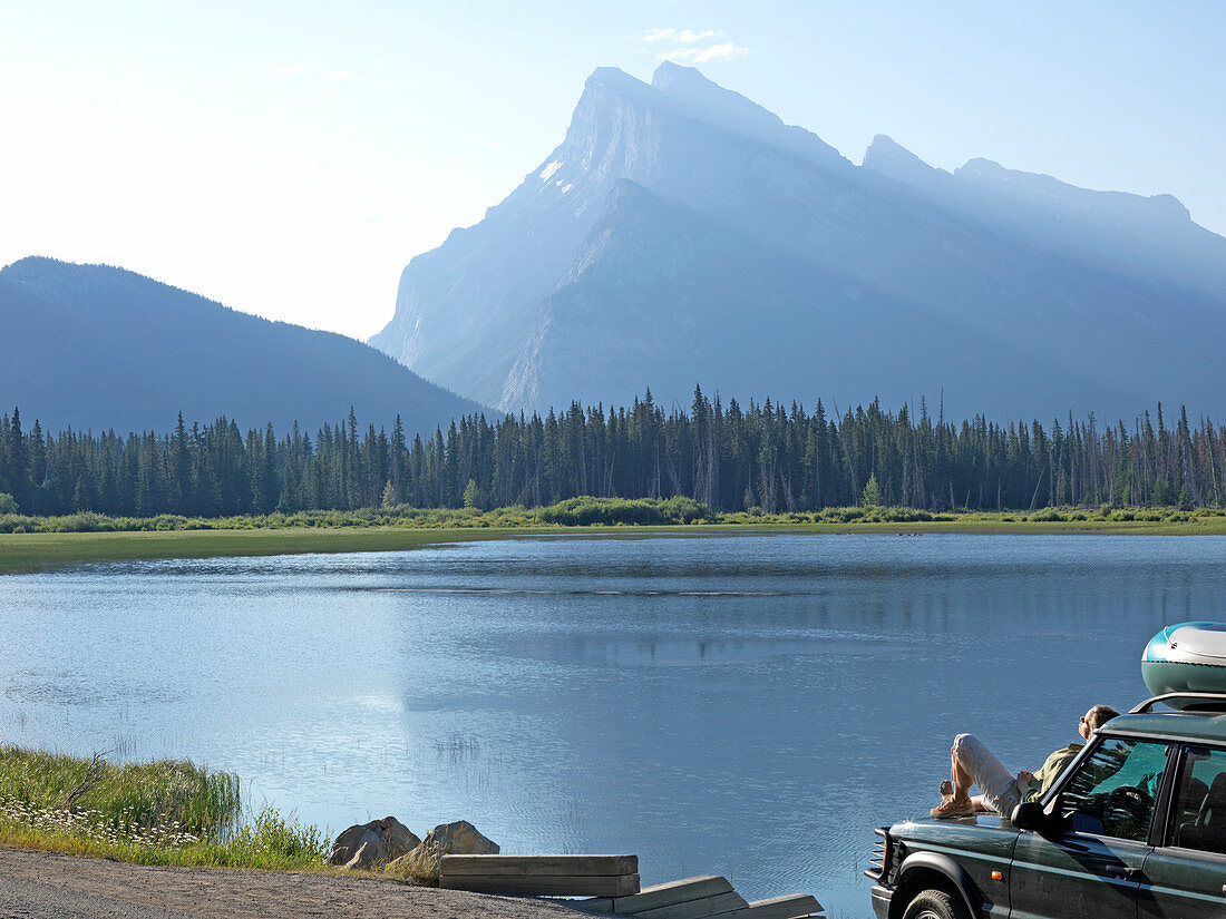 Woman relaxes on front of truck by lake and mountains