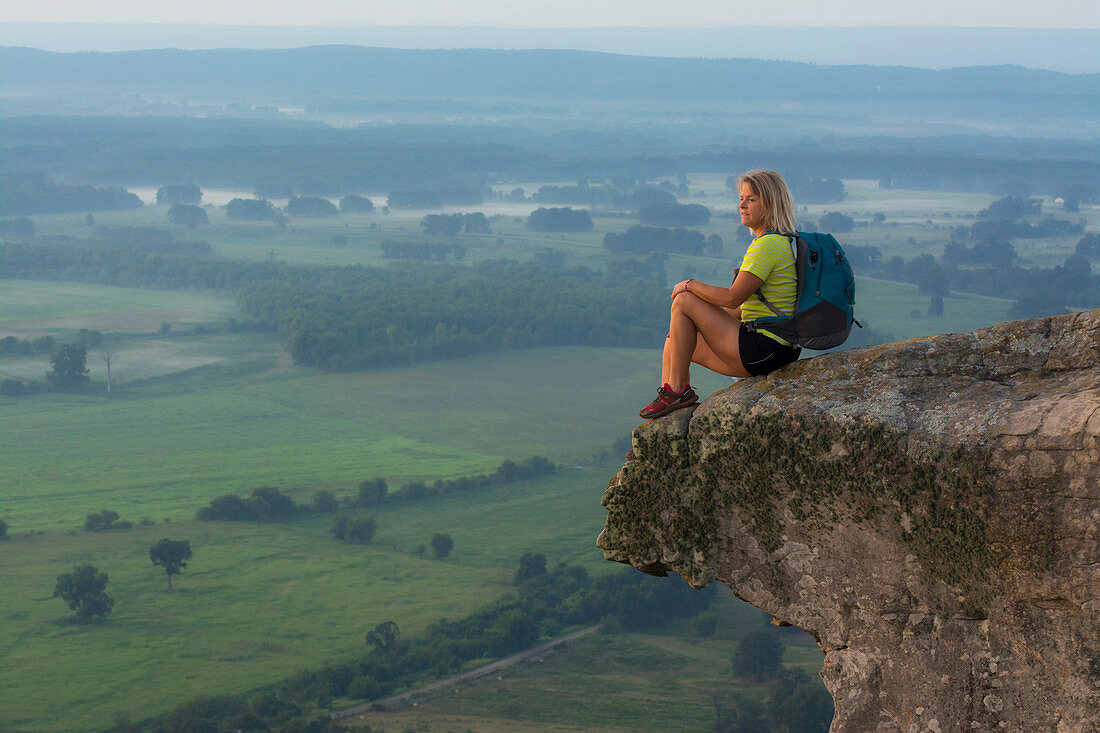 Woman sitting on sandstone overhang watching sunrise from summit of Petit Jean Mountain above Arkansas River Valley 