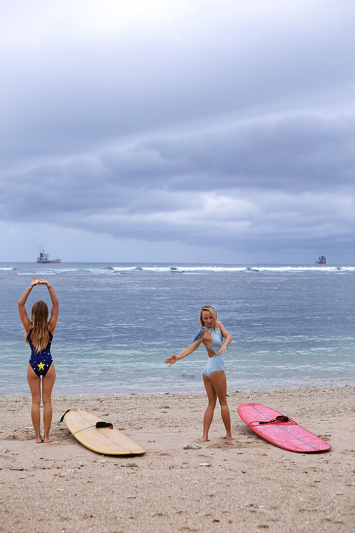 Two Young Women Stretch And Flex Before Surfing At Beach