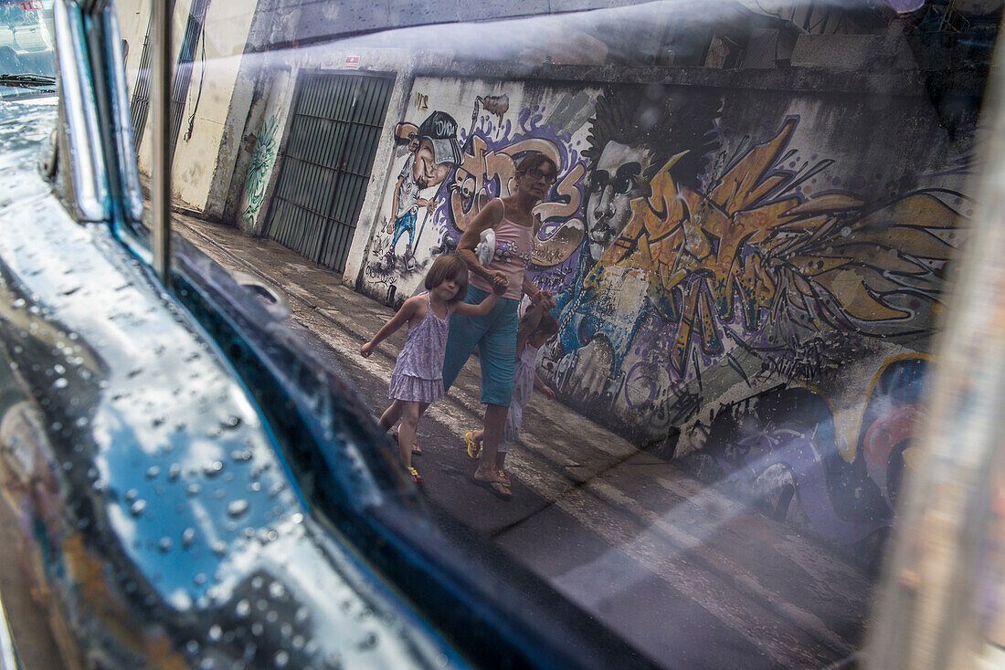 A woman walking with two young children is seen in the reflection of a window of a vintage car. A graffitied wall is behind her, and raindrops are seen on the door sill. Old Havana or La Habana Veija, La Habana, Cuba
