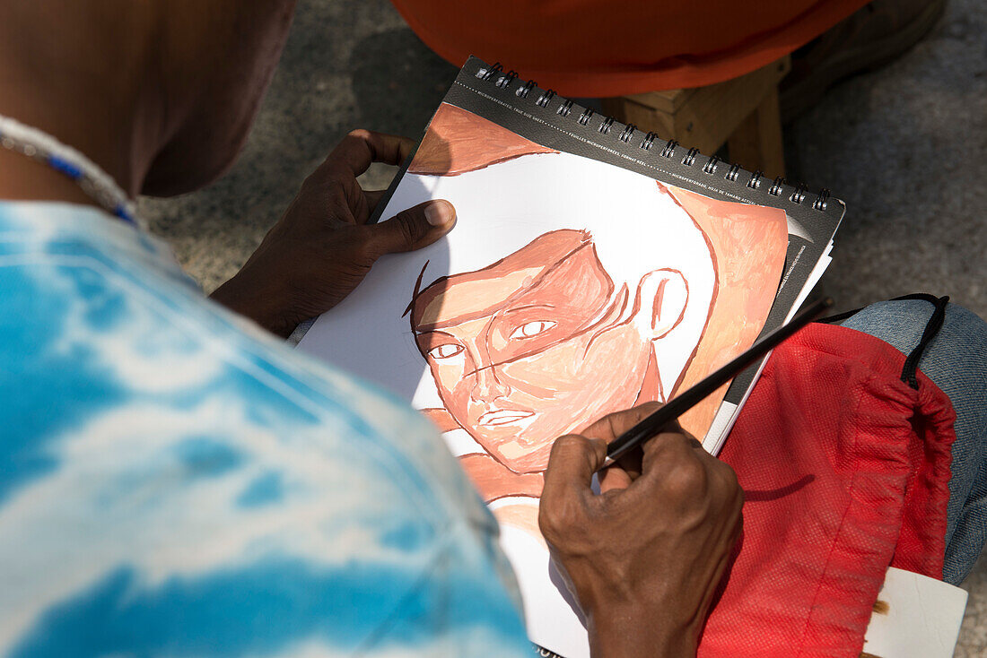 A young male Cuban street artist paints a watercolor of a girl's face on a sketchpad, as seen over his shoulder. Artist work and sell their art on El Prado Boulevard (Paeeo Marti) as families stroll on Sundays.  Centro Havana, La Habana, Cuba