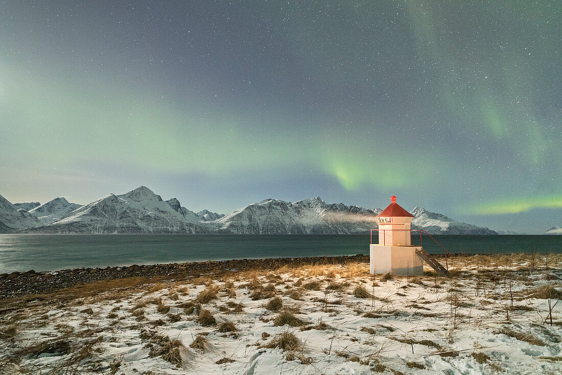 The Northern Lights (aurora borealis) and stars illuminate the lighthouse framed by icy sea, Djupvik, Lyngen Alps, Troms, Norway, Scandinavia, Europe
