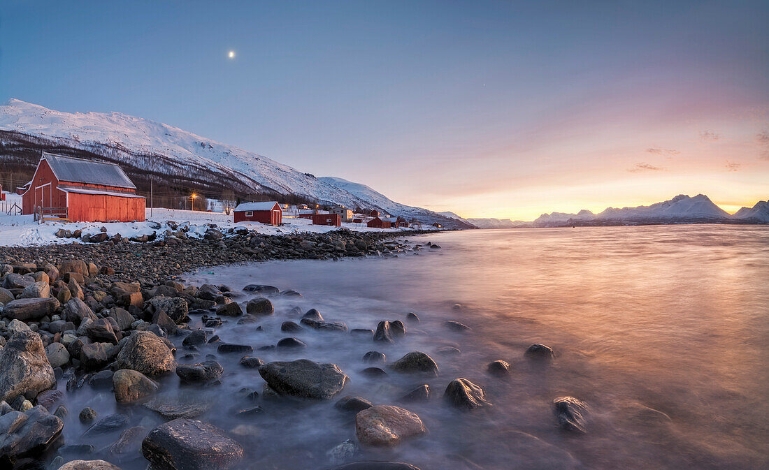 Panorama of typical wood huts called Rorbu framed by fiery sky at sunset and frozen sea, Djupvik, Lyngen Alps, Troms, Norway, Scandinavia, Europe