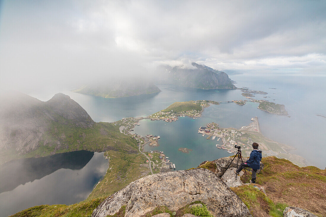 Photographer on top of rocky peak admires the blue sea surrounded by mist Reinebringen Moskenes Lofoten Islands Norway Europe