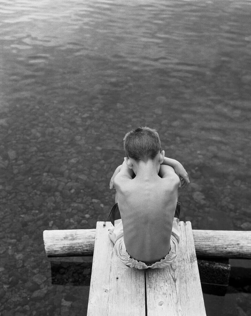 Rückansicht Porträt des traurigen Jungen Sitzen Umarmung seiner Knie am Ende des Docks am See, High Angle View