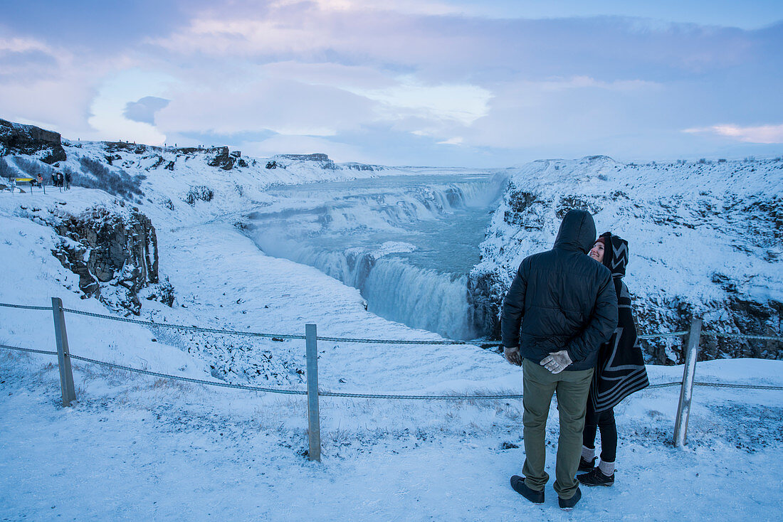 Couple overlooks the Gullfoss Falls waterfall on the river Hvítá (Hvita) in snowy landscape in winter, Gullfoss, Gullfossi, Vesturland, Iceland, Europe