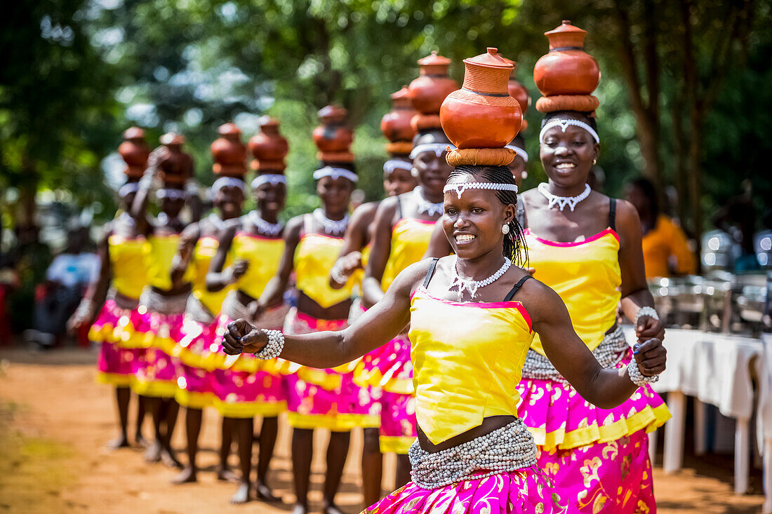 'Frauen, die hell farbige traditionelle Kleidung tragen, gehen in einer Reihe und tragen Töpferei auf ihren Köpfen; Uganda'
