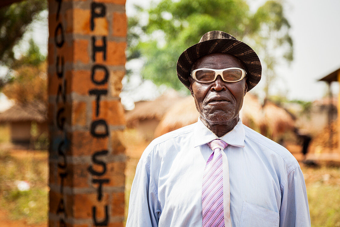 'A man with white eyeglasses stands posing outside a photo studio; Uganda'