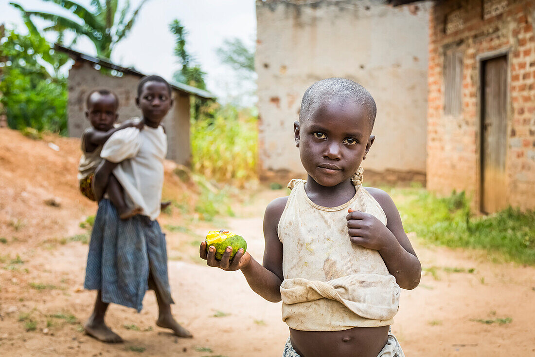 'A young child stands holding a piece of fruit with a girl holding a young child on her back in the background; Uganda'