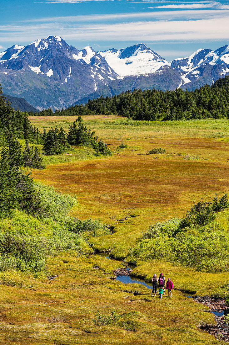 'A small group of women hiking along a stream in Turnagain Pass, with patches of snow on the Chugach Mountains in the background; Alaska, United States of America'