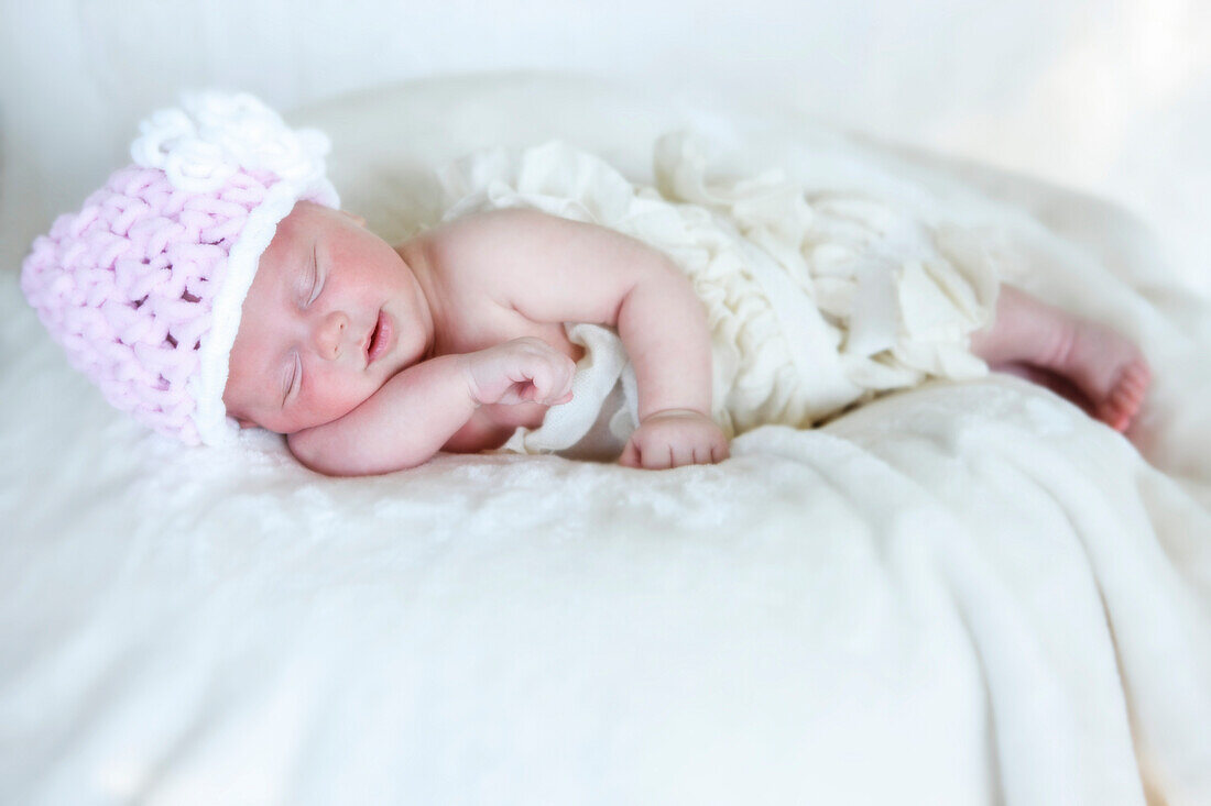 'A newborn baby lays sleeping on a white blanket wearing a pink knit hat; Washington, United States of America'