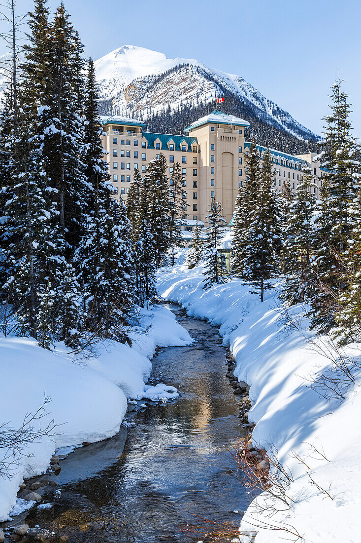 'Winter trails along Lake Louise in Banff National Park, the river is not fully frozen at the edge of the Fairmont Chateau Lake Louise Resort shown against the mountains in the background; Lake Louise, Alberta, Canada'