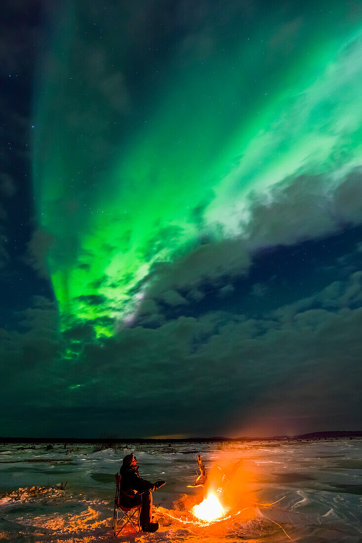 Ein Mann erwärmt sich neben einem Treibholzfeuer, während er die Aurora durch Wolken am gefrorenen Delta River, in der Nähe von Delta Junction, Alaska, beobachtet.