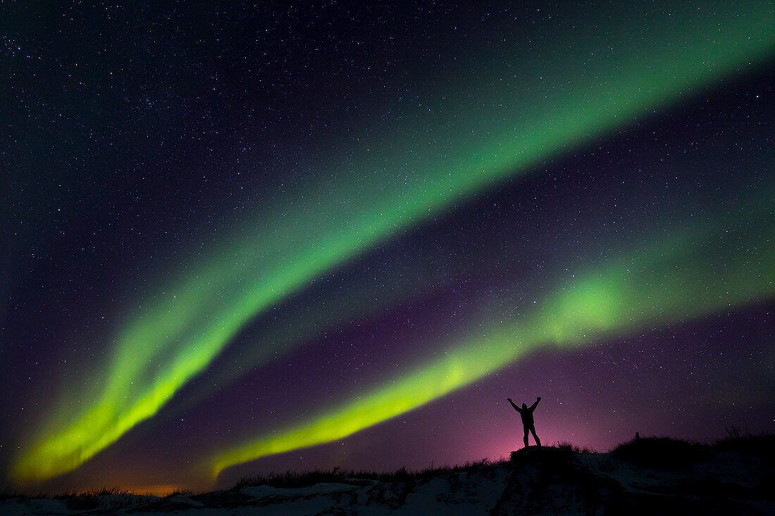 'Colourful Aurora borealis over a man with arms outstretched silhouetted against light pollution from nearby Fort Greely; Alaska, United States of America'