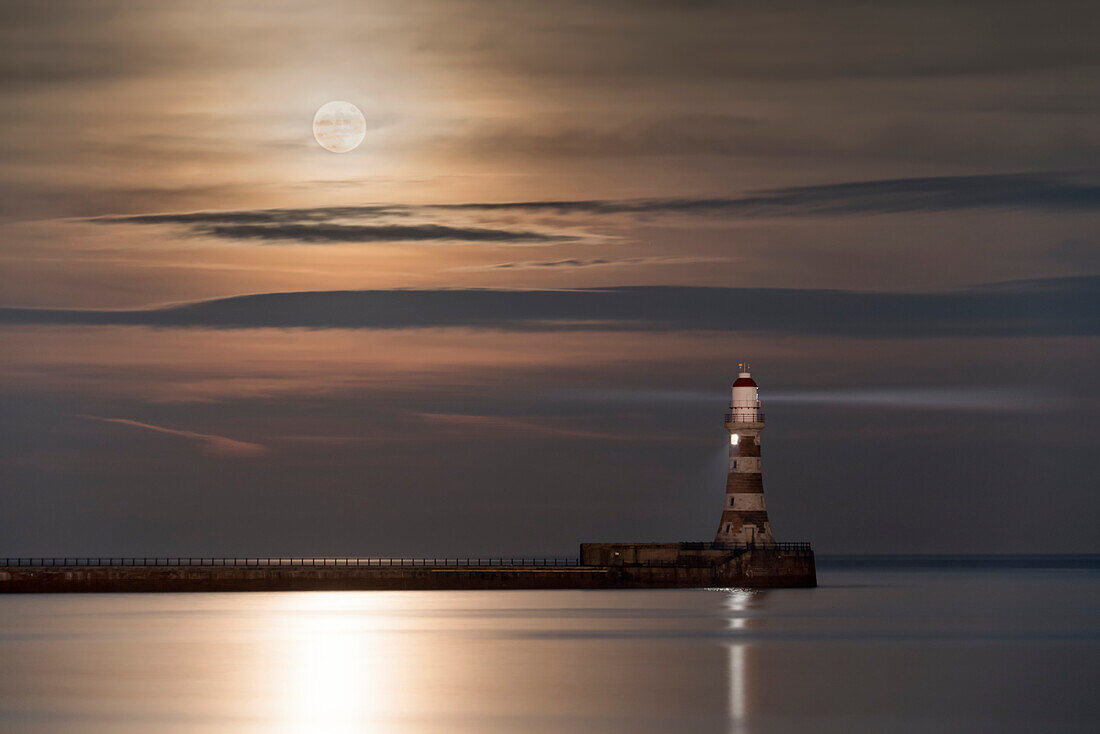 Roker Lighthouse glühend am Ende eines Pier unter einem hellen Vollmond, der auf ruhigem Wasser reflektiert, Sunderland, Tyne und Wear, England