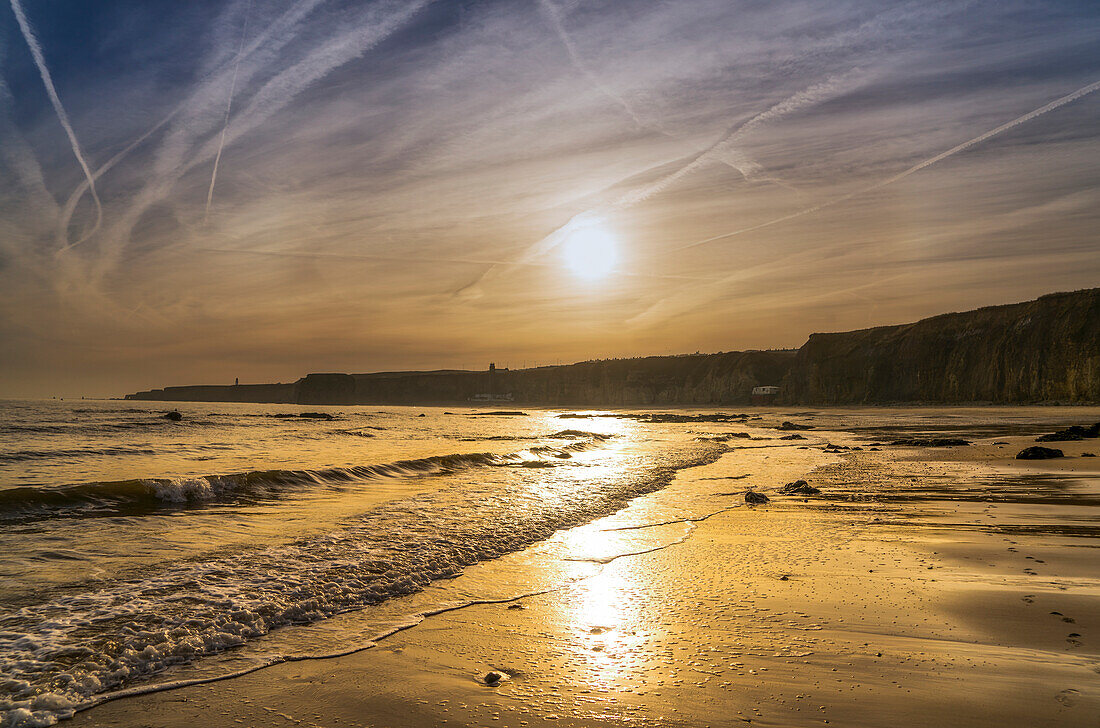 Eine sinkende Sonne leuchtet in den Himmel und reflektiert den nassen Sand entlang der Küste, South Shields, Tyne und Wear, England