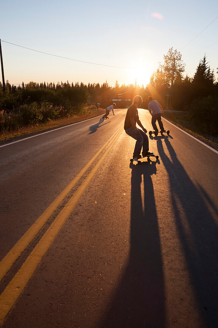 Three young men skateboarding down a road at sunset, Homer, Alaska, United States of America