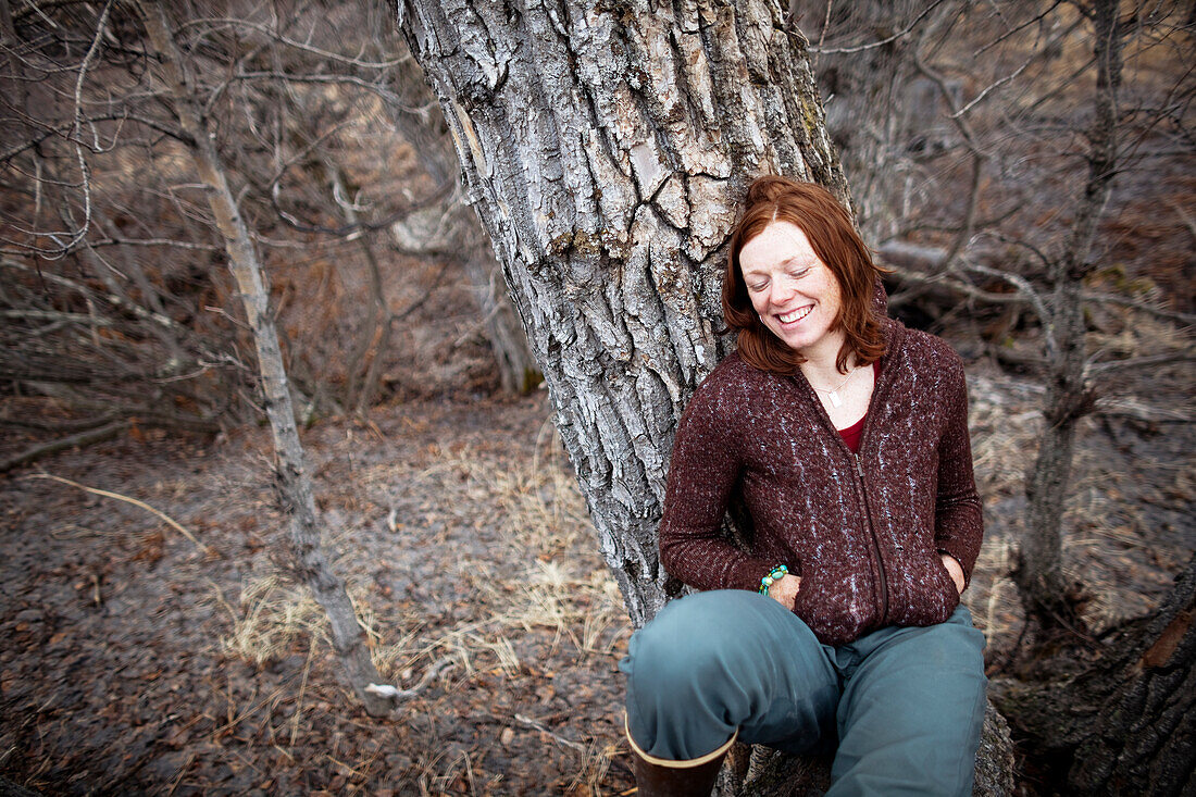 Portrait of a woman with red hair leaning on a tree and smiling with eyes closed, Homer, Alaska, United States of America