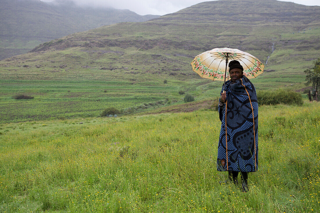 Ein Mann hält einen Regenschirm und in eine blaue Decke gehüllt steht in den Bergen, Lesotho, Afrika