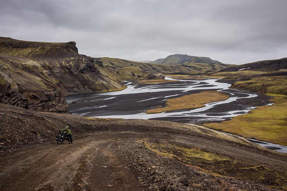 Woman dressed in waterproof equipment with a helmet on, pushing a loaded touring bicycle up a dirt/ gravel road in an inhospitable landscape