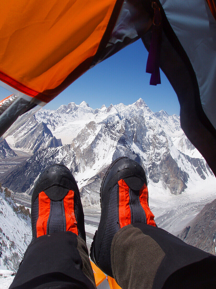 Blick auf die Karakoram Berge von Pakistan aus einem Zelt hoch an den Hängen des Broad Peak, einer der Welten 8000 Meter hohen Gipfeln. Die Expeditionsstiefel eines Bergsteigers ragen aus dem Vestibül heraus, während sie sich ausruhen.