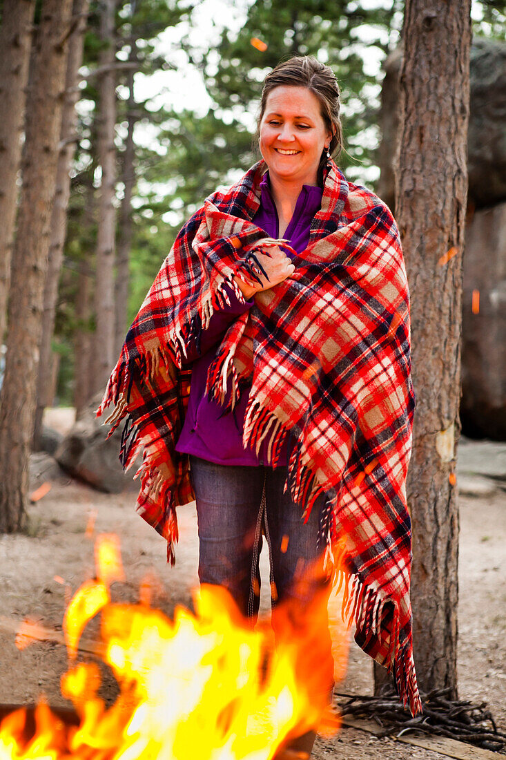 A woman smiles while she stands next to a campfire with a plaid wool blanket wrapped around her.