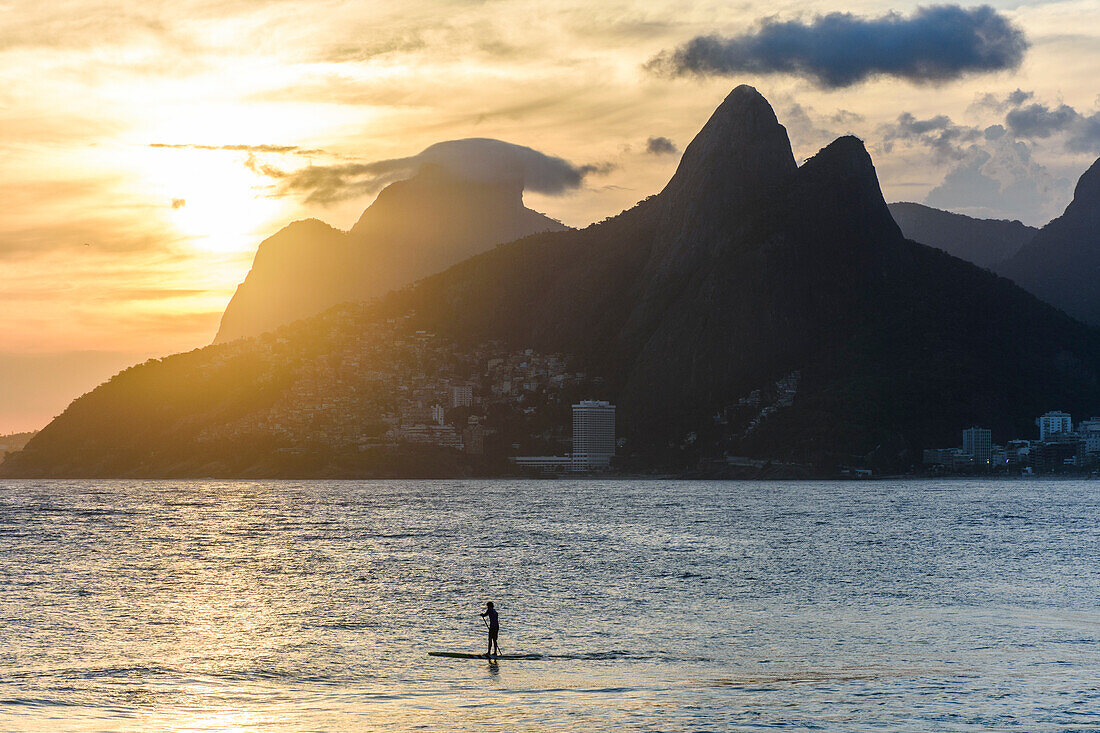 Local riding a stand up paddle board during sunset in Ipanema Beach, Morro Dois Irmaos on the back, Rio de Janeiro, Brazil