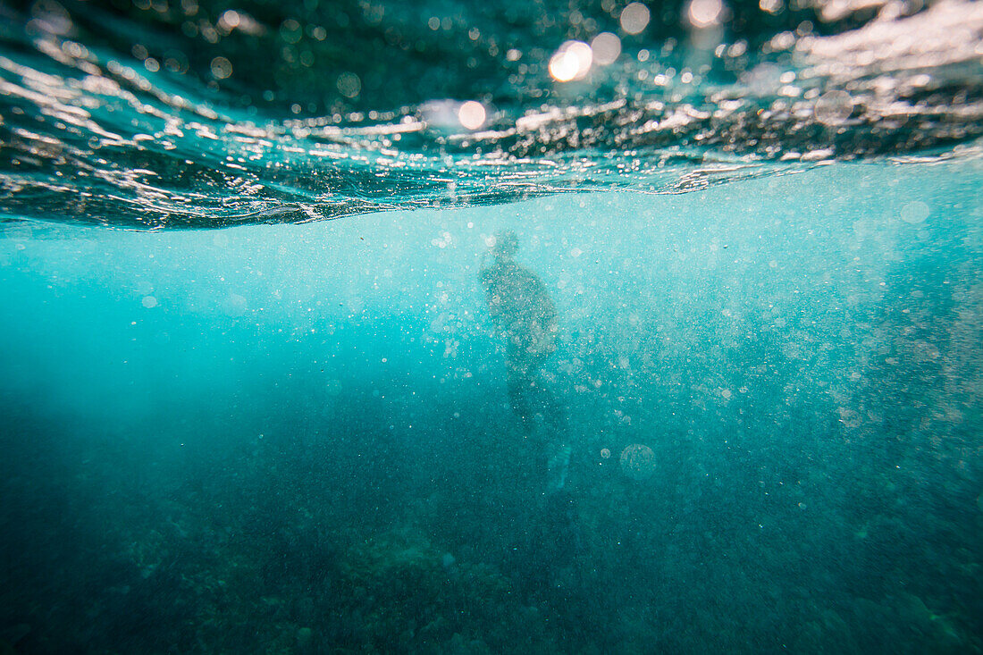 Der Umriss eines Schnorchlers wird durch Blasen und turbulente Strömungen unter Wasser gesehen, während Wellen auf Felsen und Riff vor Utila Insel, Honduras brechen.