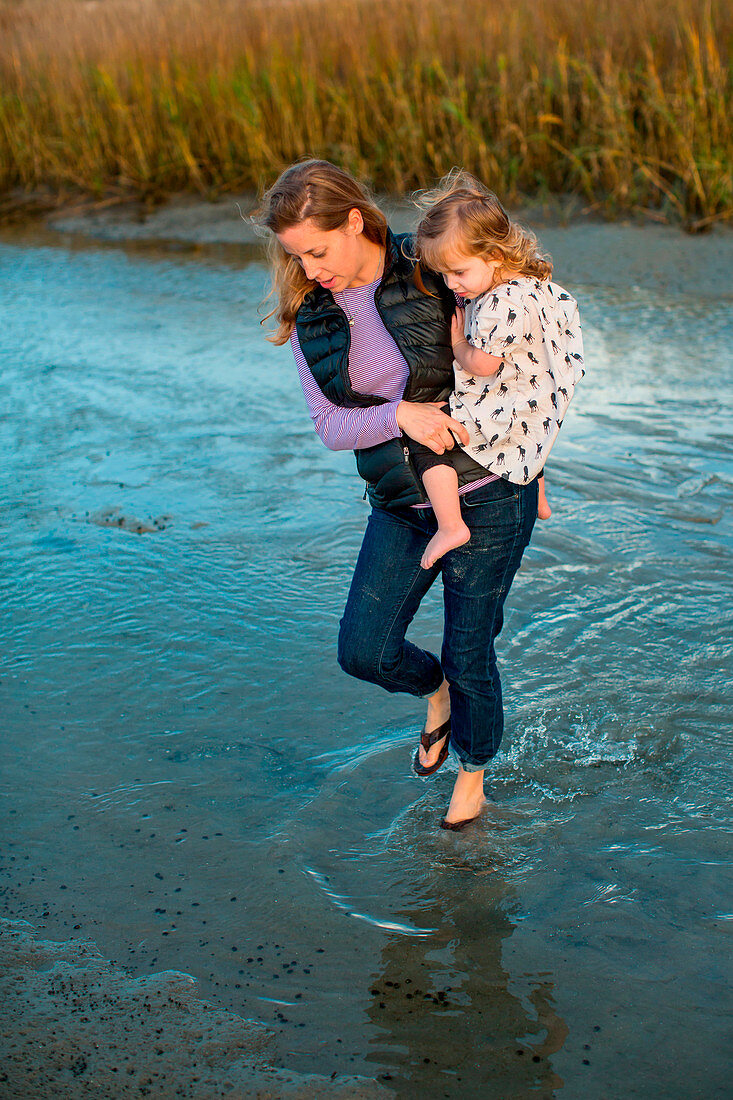 A woman walks through shallow water carrying her daughter in her arms in a marsh at Wrightsville Beach, North Carolina.