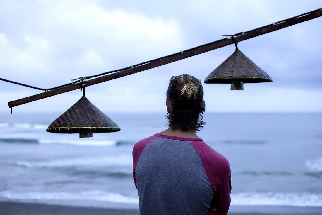 Rear view of man looking at ocean beside light fixtures hanging outdoors, Bali, Indonesia