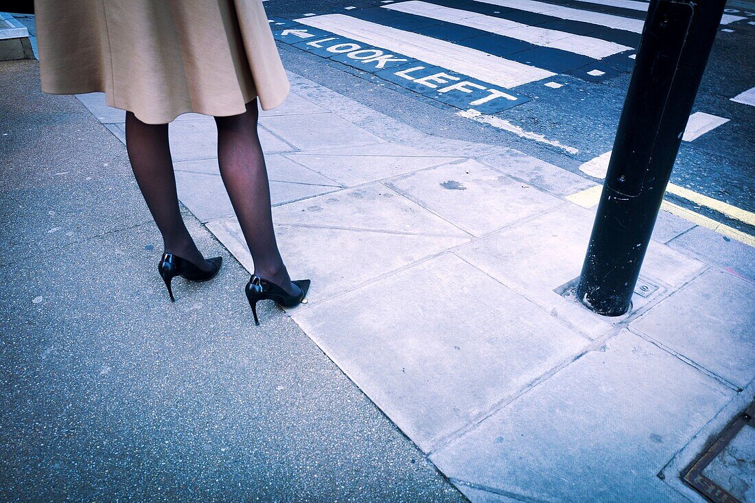 Close-up der Beine einer Frau in Mode mit einem Mantel, schwarzen Strümpfen und High Heels in der Straße neben Fußgängerüberweg gekleidet. London, England