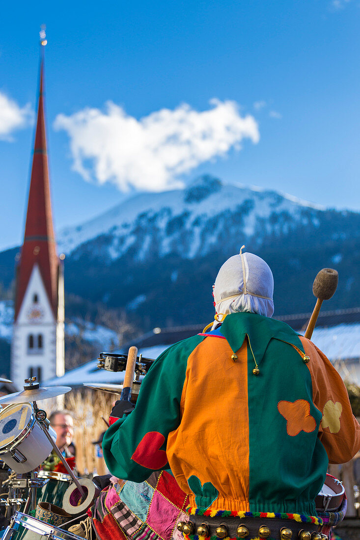 Während des Karnevals durch die Straßen des Dorfes spielen maskierte Figuren mit ihren Instrumenten, im Hintergrund die Turmglocke von Axams, Axamer Wampelerreiten, Axams, Inntal, Tirol, Osterreich (Österreich), Europa