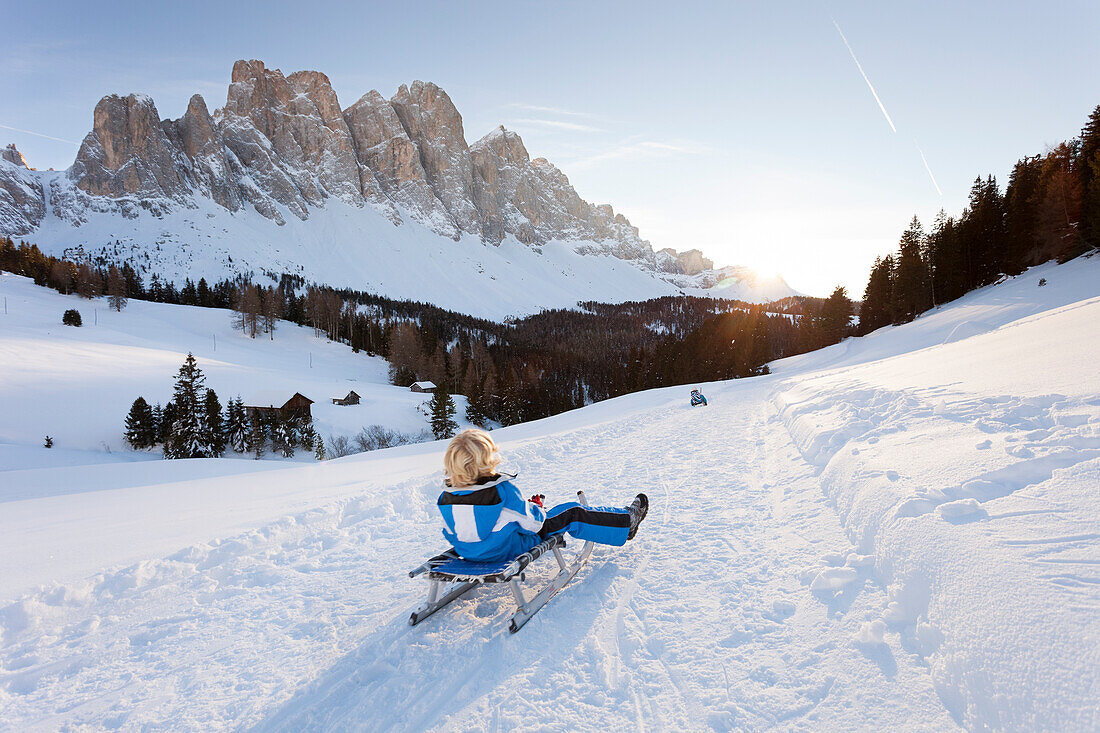 Ein Kind geht mit einem Schlitten bei einem schönen Sonnenuntergang in Villnoss mit dem Geisler im Hintergrund, Provinz Bozen, Südtirol, Trentino-Südtirol, Italien, Europa